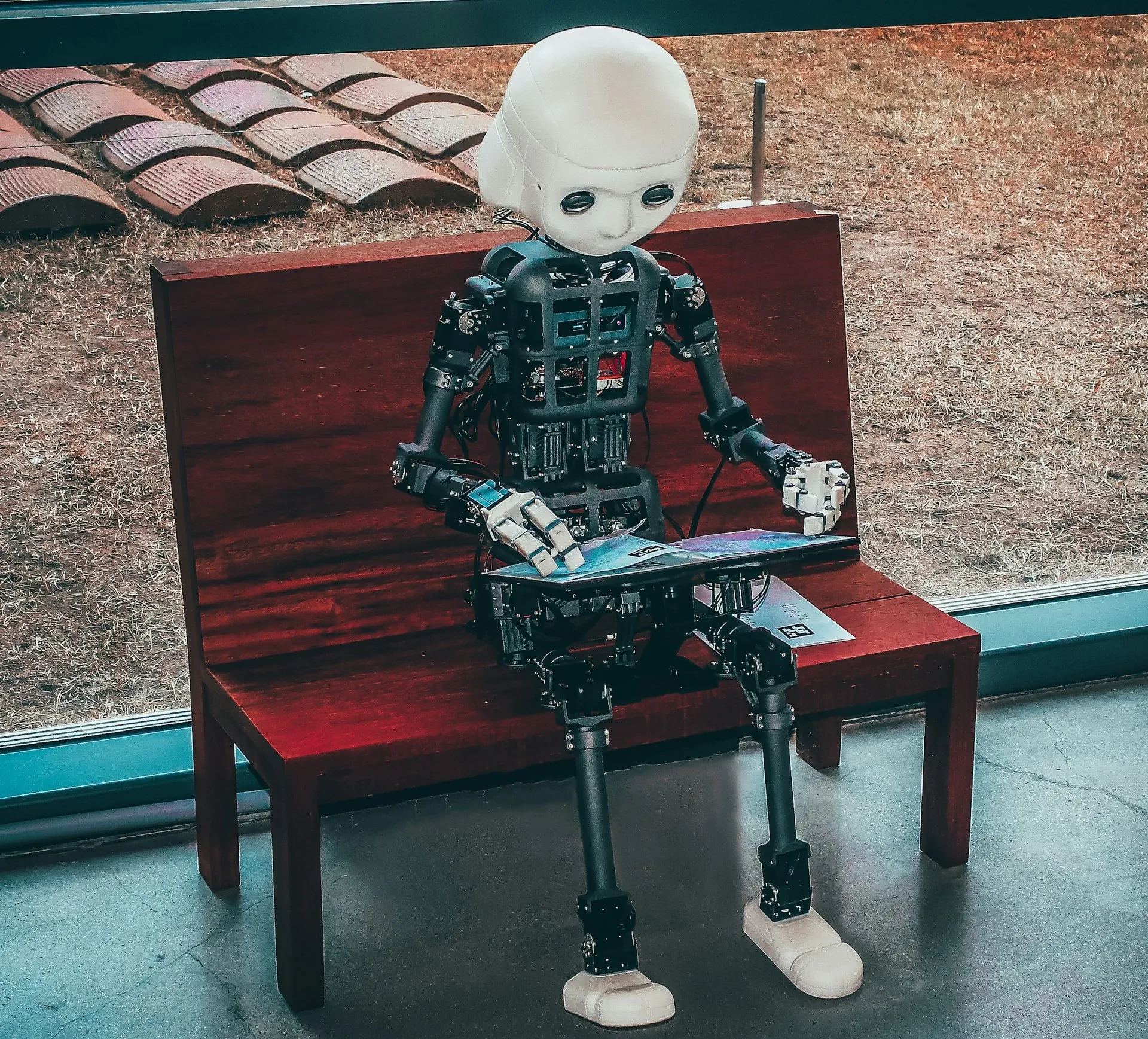 Humanoid robot sitting on a bench, reading a magazine, with a white head, mechanical limbs, and an exposed framework, set against a glass window background.