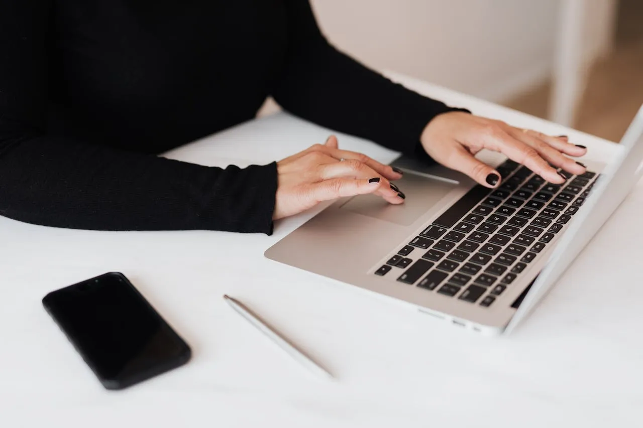 Woman typing on a laptop with a smartphone and pen nearby.