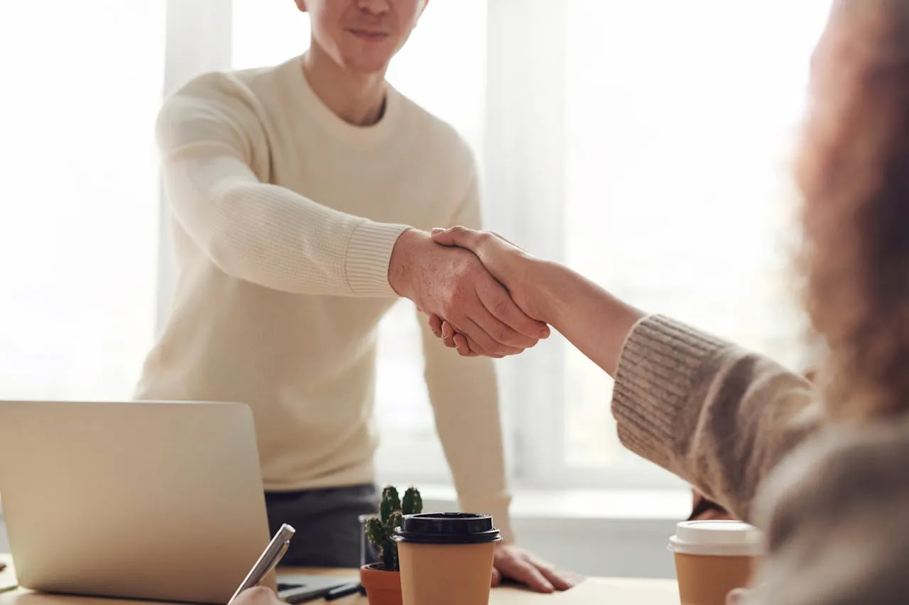 Two professionals shaking hands in office with laptop and coffee.